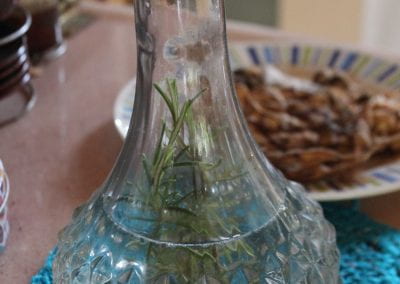 On a table indoors in Tuwaliri's cloud forest, a small, thin plant sits suspended in water inside of a crystalline vase, which sits on top of a blue table mat next to a plate of another dried plant