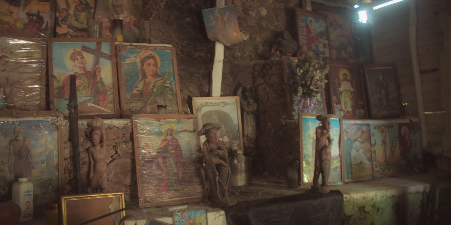 Large framed images lie against a rocky natural wall, arranged among candles and small statues.