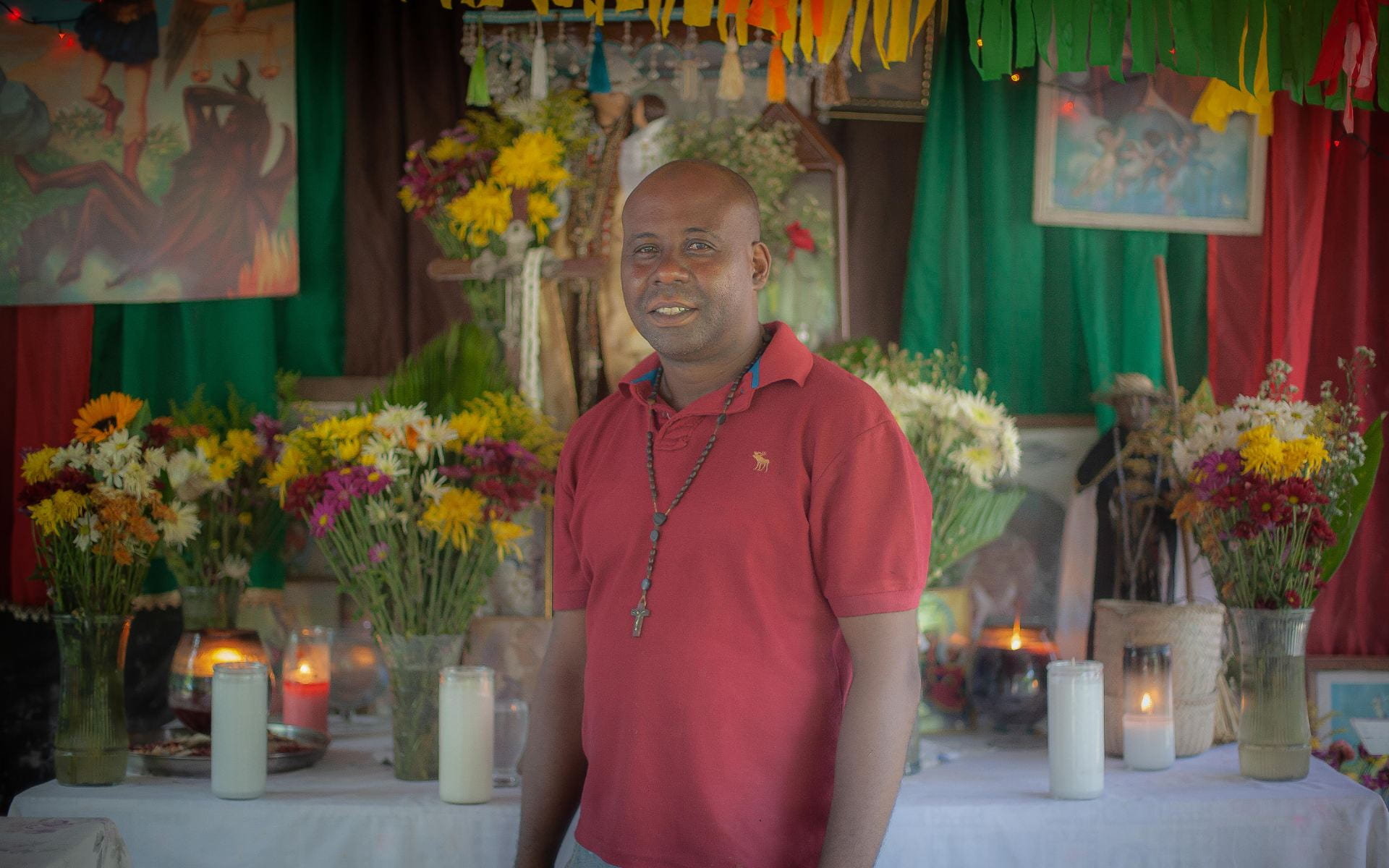 Ramoncito stands and looks into the camera in front of his altar laden with flowers and candles, wearing a red polo and rosary necklace.