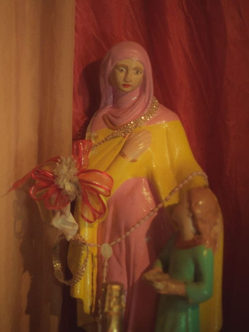 A statue of a feminine figure wearing a pink hood and yellow dress, with one arm holding flowers and the other arm wrapped around a small child.