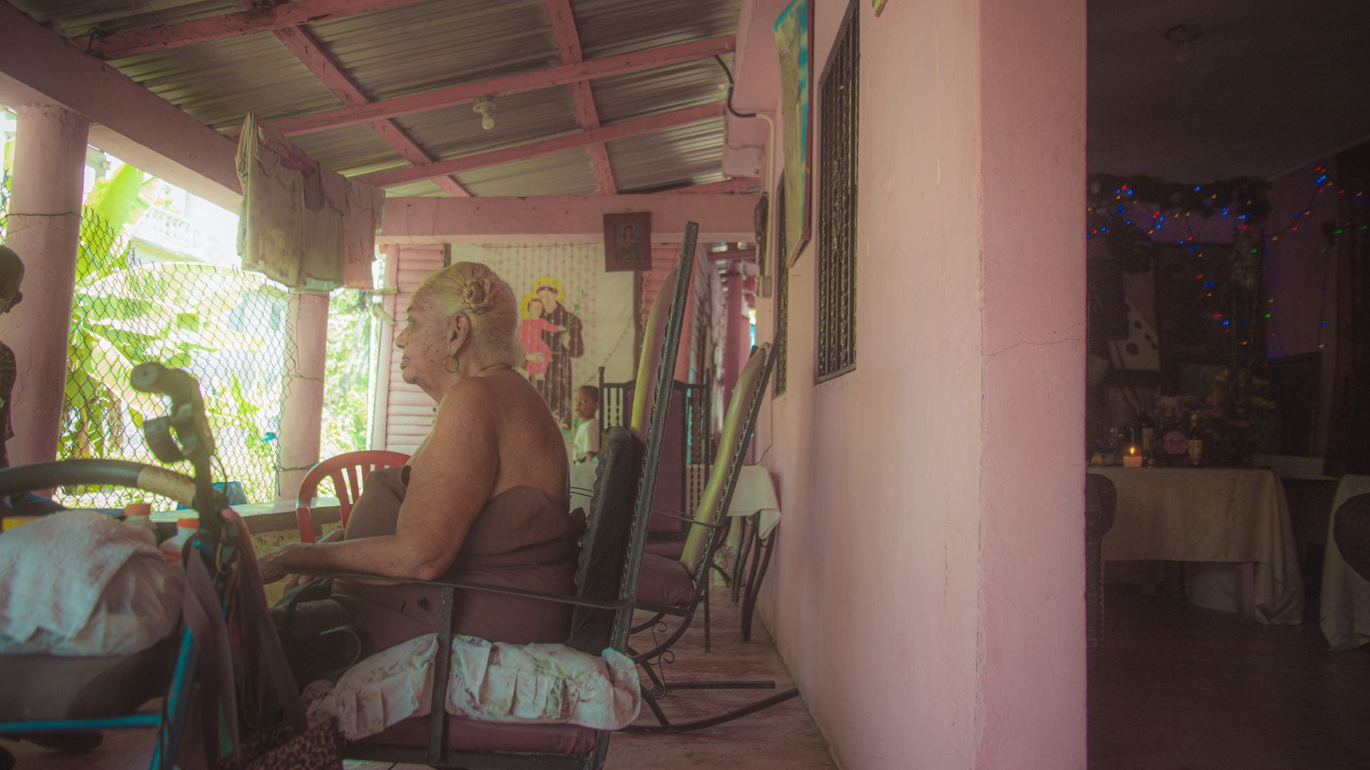 Graciela sits at a table facing away from the camera, outside of her home with bright pink walls.